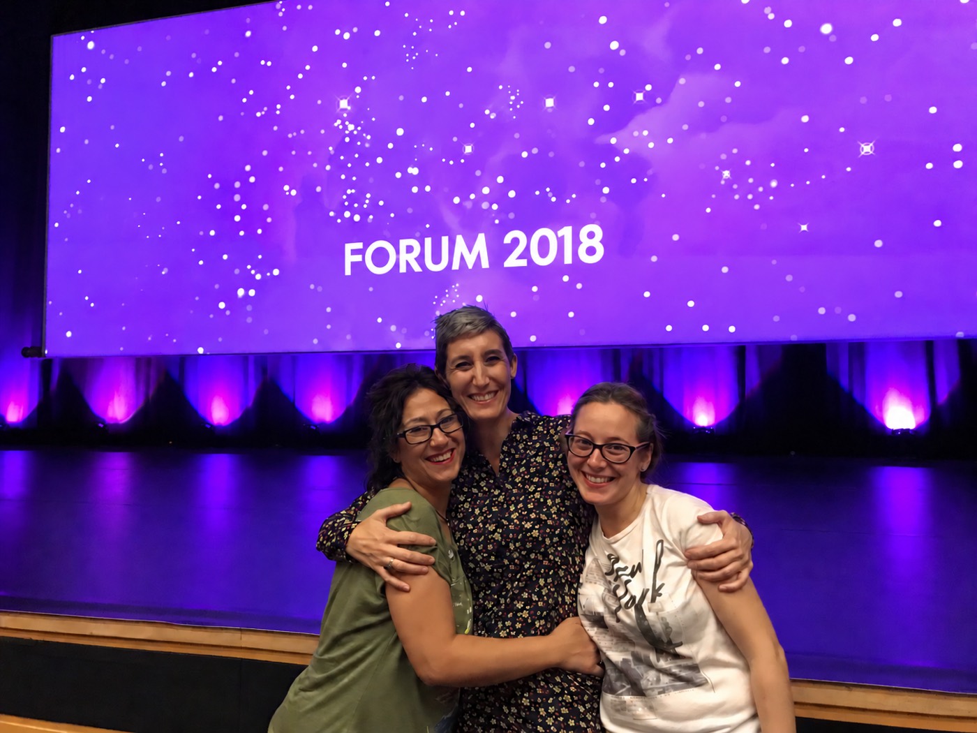 Irene Molina con Pepi Rodríguez y Encarni López, abrazadas y sonrientes en el escenario del Fórum 2018 en Valencia, frente a una pantalla morada