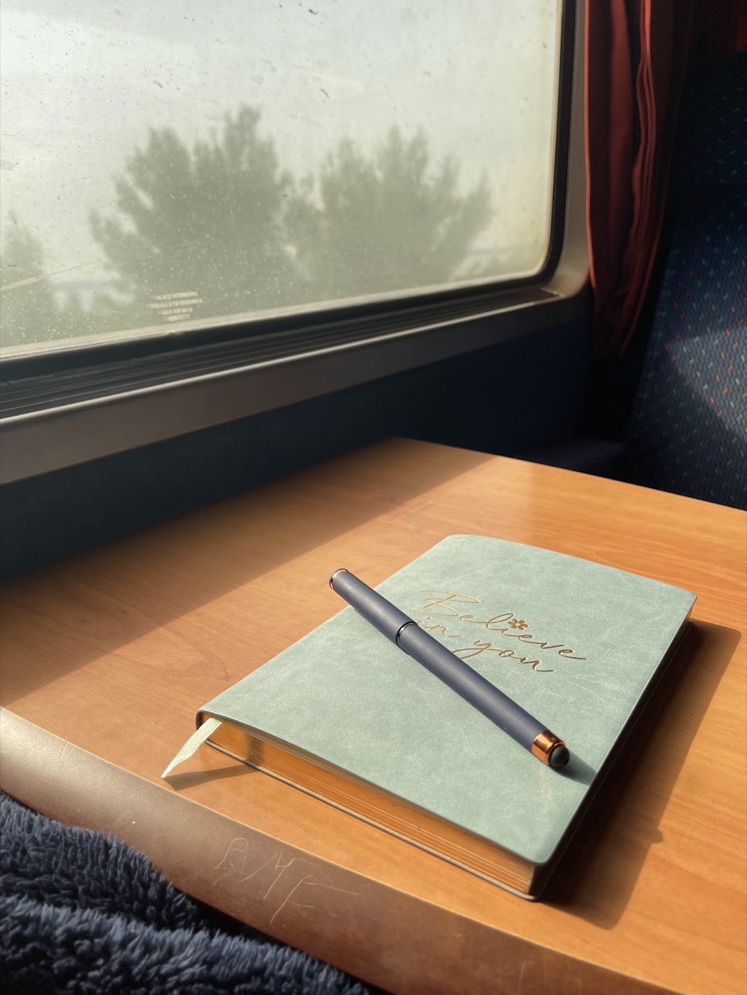 Cuaderno verde con una pluma sobre la mesa junto a la ventanilla de un tren, iluminado por la luz del sol