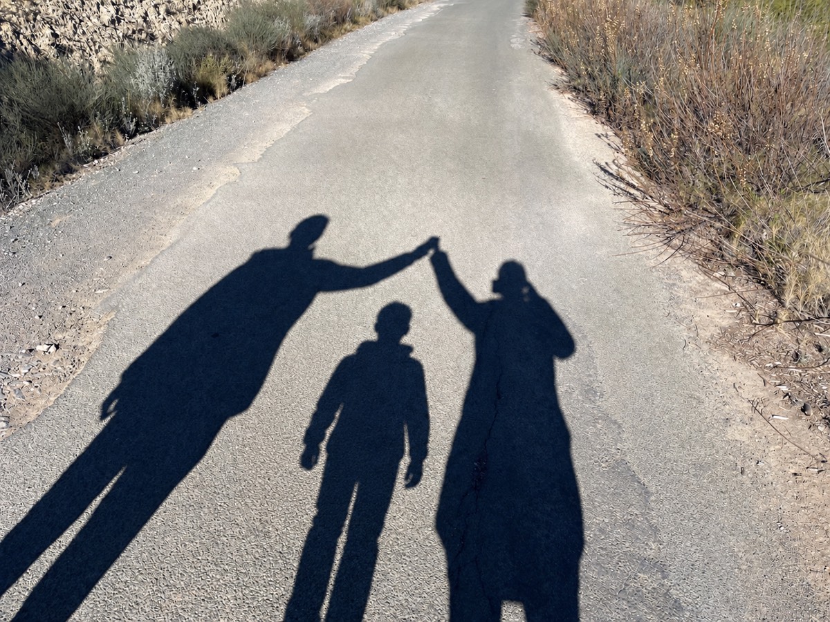 Sombras de una familia sobre un camino: dos adultos con las manos en alto y un niño entre ellos